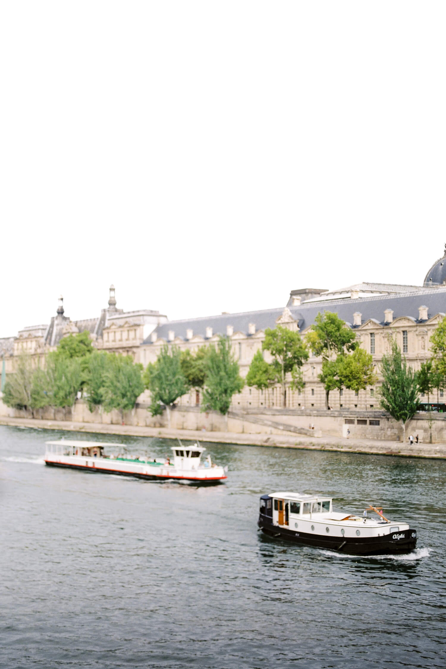 two boats cruising on the Seine River. The background is showing the exterior or the iconic Louvre Museum and the riverbank viewed from Île de la Cité. Shot on analogue film by Paris wedding photographer Cristina Ilao